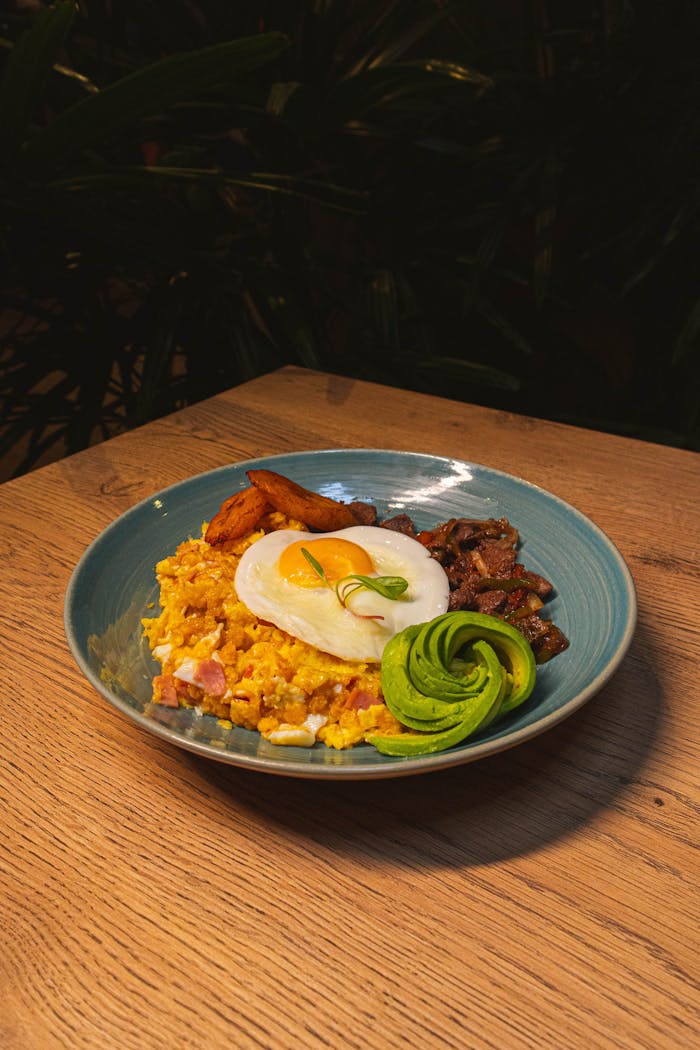 about-us A vibrant breakfast plate features fried egg, avocado, plantain, and rice on a wooden table.